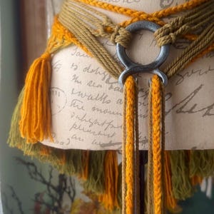 May include: A brown and yellow braided belt with a metal ring buckle and tassels. The belt is tied around a mannequin and the tassels are hanging down.