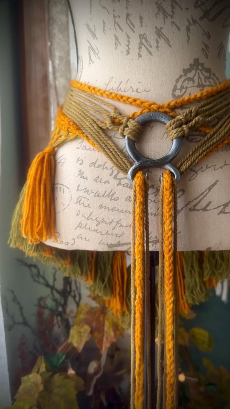 May include: A brown and yellow braided belt with a metal ring buckle and tassels. The belt is tied around a mannequin and the tassels are hanging down.