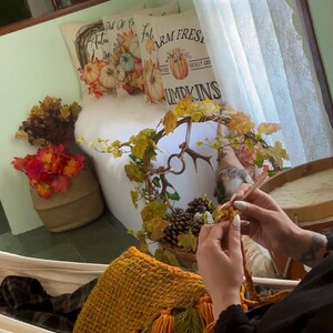 May include: A person sits in a white hammock with a yellow and green woven bag. The person is knitting with a white yarn. The background shows a white couch with two pillows with fall-themed designs. The pillows have the text "Fall" and "Farm Fresh Locally Grown Pumpkins".