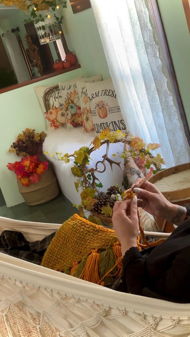 May include: A person sits in a white hammock with a yellow and green woven bag. The person is knitting with a white yarn. The background shows a white couch with two pillows with fall-themed designs. The pillows have the text "Fall" and "Farm Fresh Locally Grown Pumpkins".