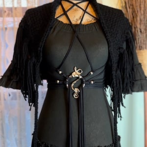 Könnte beinhalten: Ein schwarzes Kleid mit einem silberfarbenen Gürtel und einem passenden schwarzen Halsband. Ein schwarzer Fransen-Schal ist über den Schultern drapiert. Das Kleid hat einen gerüschten Rock und ein dekoratives Geschirr.