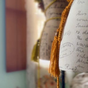 May include: A close-up of a vintage-style dress form with a handwritten note on the fabric. The dress form is decorated with a long, orange tassel and a shorter, yellow tassel. The tassels are made of yarn and have a knotted fringe.