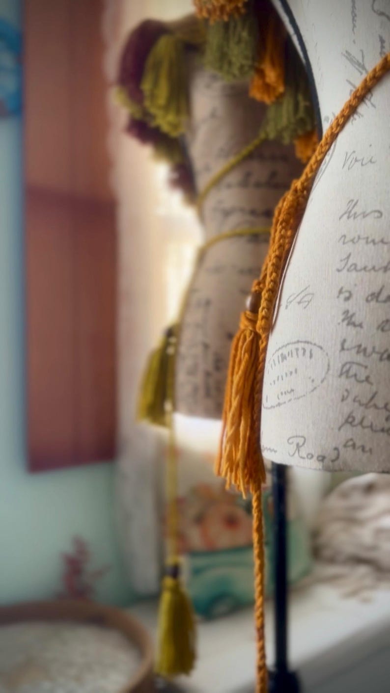 May include: A close-up of a vintage-style dress form with a handwritten note on the fabric. The dress form is decorated with a long, orange tassel and a shorter, yellow tassel. The tassels are made of yarn and have a knotted fringe.