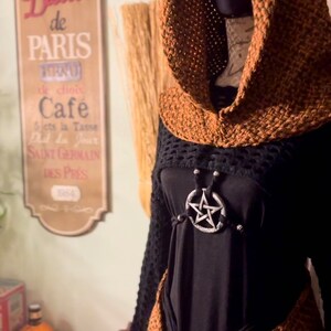 May include: A brown crocheted hooded top with fringe trim and a black mesh top underneath. The top is worn over a black shirt with a silver pentagram pendant.