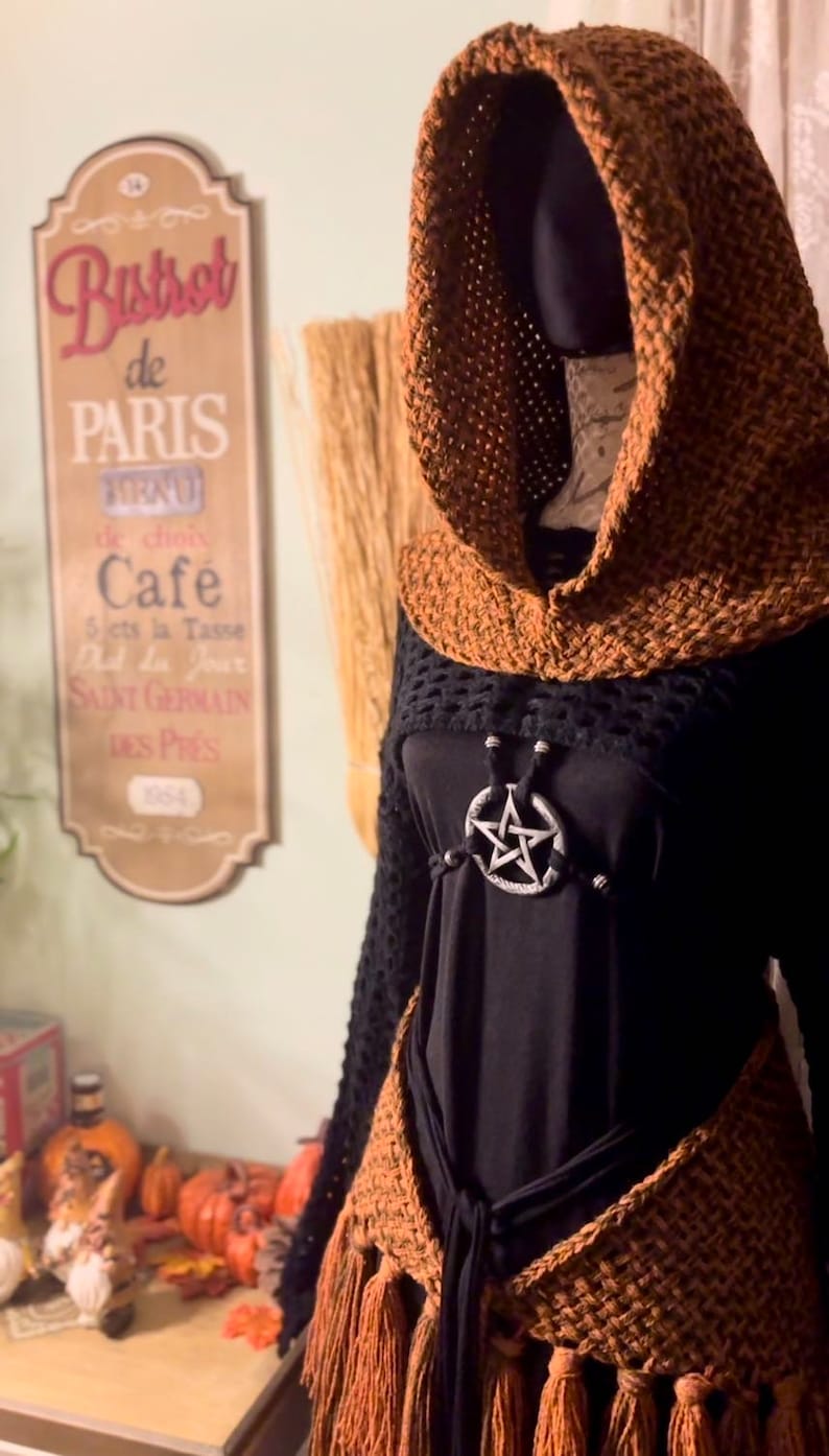 May include: A brown crocheted hooded top with fringe trim and a black mesh top underneath. The top is worn over a black shirt with a silver pentagram pendant.