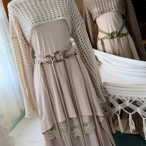 May include: Two beige crochet tops with long sleeves and a loose fit. The tops are layered over beige dresses with lace trim and long fringe belts.
