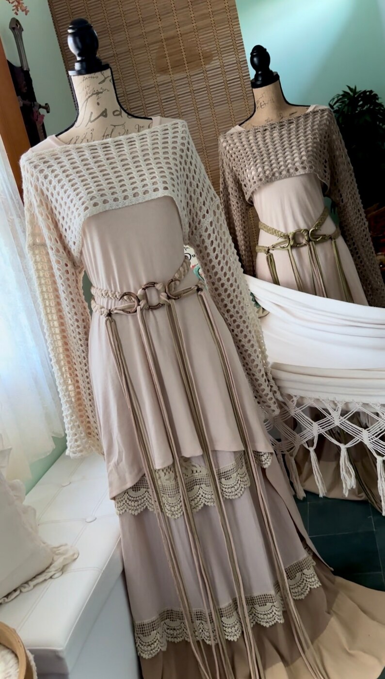 May include: Two beige crochet tops with long sleeves and a loose fit. The tops are layered over beige dresses with lace trim and long fringe belts.