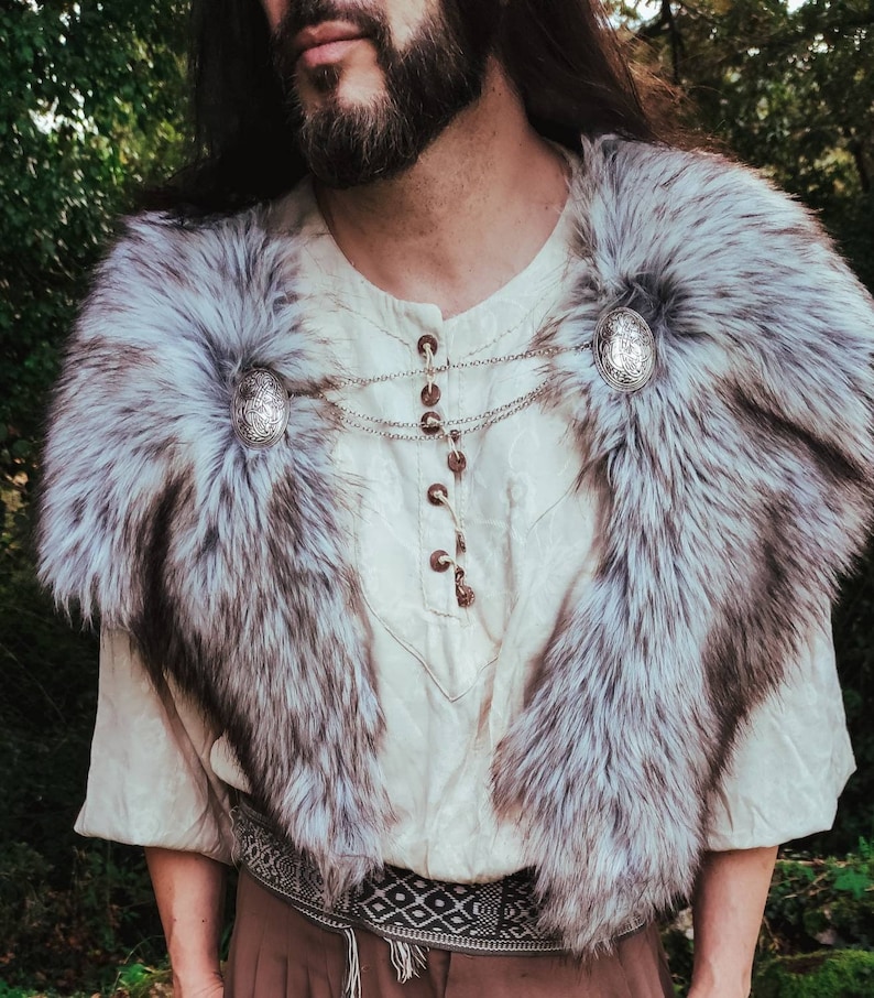May include: A white faux fur collar with silver accents. The collar is worn over a white shirt with a brown button-up closure. The shirt has a geometric pattern belt.