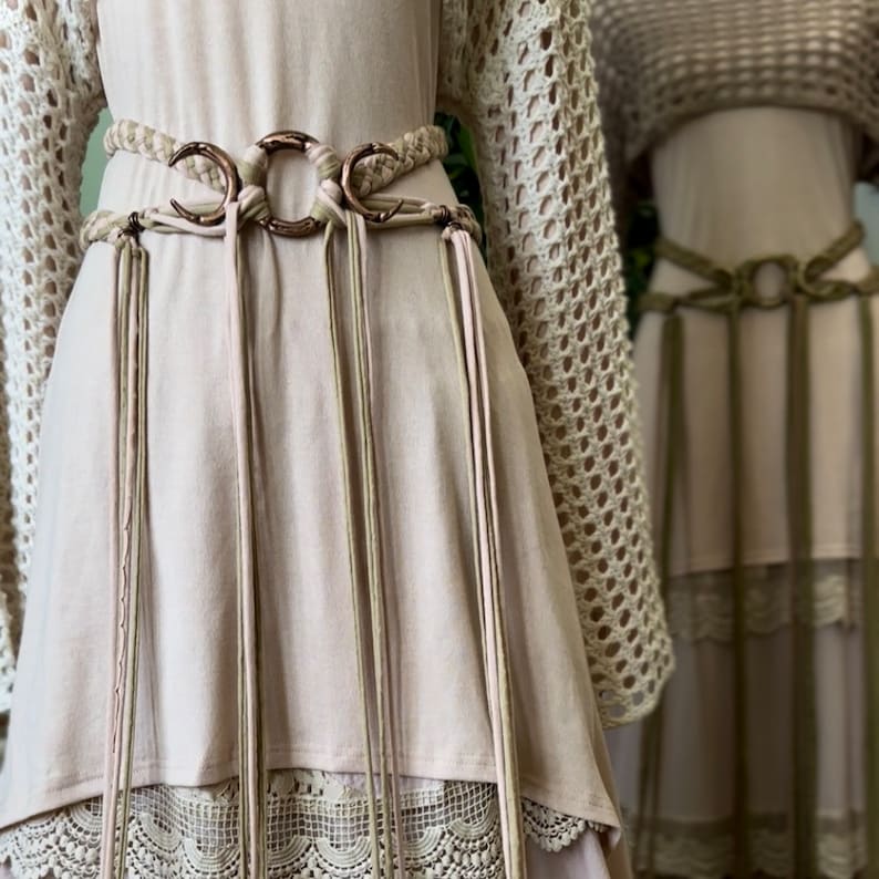 May include: A light pink dress with a crocheted lace trim and a brown leather belt with crescent moon shaped buckles and tassels.