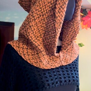 May include: A brown knitted hooded scarf with a woven pattern. The scarf is worn on a black shirt with a crocheted design.