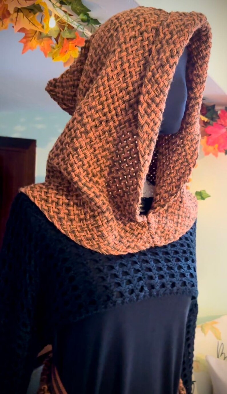 May include: A brown knitted hooded scarf with a woven pattern. The scarf is worn on a black shirt with a crocheted design.