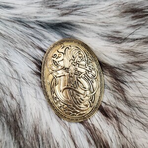 K&ouml;nnte beinhalten: Eine ovale Brosche in Goldfarbe mit einem komplizierten Design aus ineinandergreifenden Linien und Formen. Die Brosche ist vor einem wei&szlig;en und grauen Kunstfellhintergrund platziert.