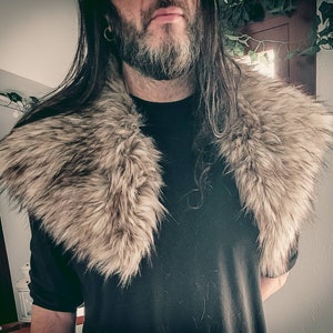 K&ouml;nnte beinhalten: Eine Person tr&auml;gt ein schwarzes Shirt mit einem gro&szlig;en, flauschigen, grauen Pelzkragen. Der Kragen ist dick und bedeckt den Hals und die Schultern der Person.