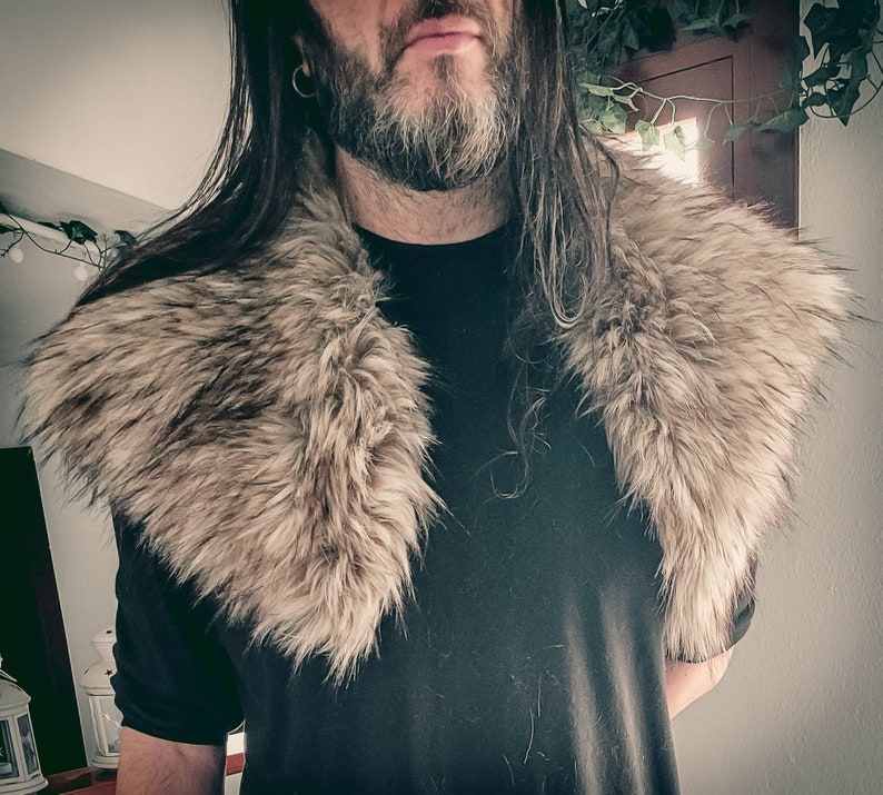 K&ouml;nnte beinhalten: Eine Person tr&auml;gt ein schwarzes Shirt mit einem gro&szlig;en, flauschigen, grauen Pelzkragen. Der Kragen ist dick und bedeckt den Hals und die Schultern der Person.