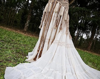 May include: A white bohemian-style wedding dress on a mannequin. The dress features a lace bodice, a fringed belt, and a flowing skirt. A faux fur shawl is draped over the shoulders. The dress is displayed outdoors in a grassy area with trees in the background.