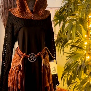 May include: A brown crocheted hooded top with fringe and a black long-sleeved shirt underneath. The top is worn with a brown leather belt with a pentagram buckle and brown antler accents.