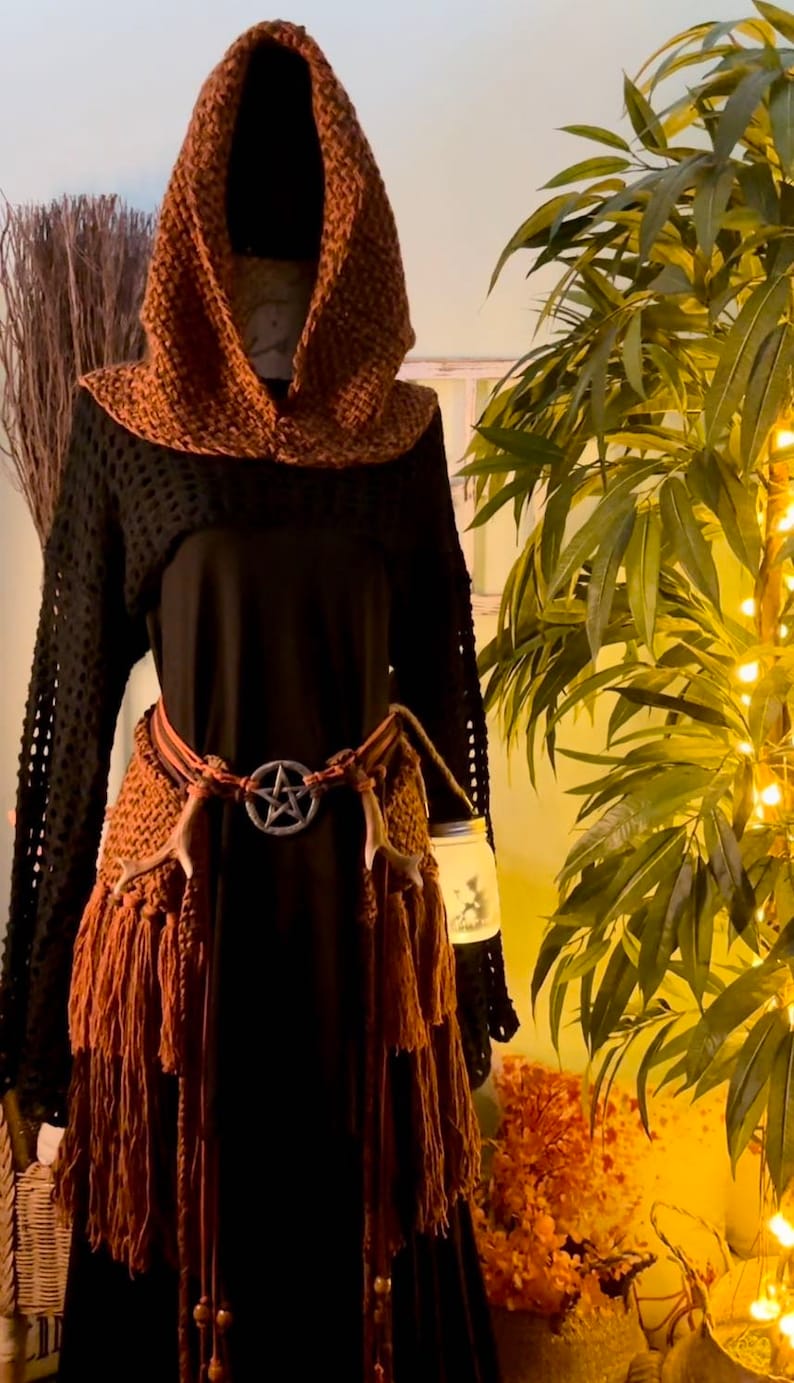 May include: A brown crocheted hooded top with fringe and a black long-sleeved shirt underneath. The top is worn with a brown leather belt with a pentagram buckle and brown antler accents.