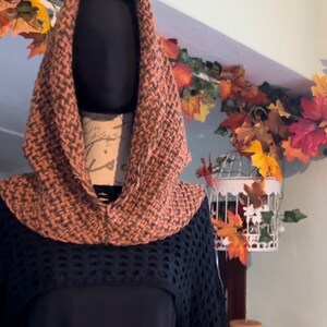 May include: A brown and orange knitted cowl with a hood. The cowl is displayed on a mannequin wearing a black top.