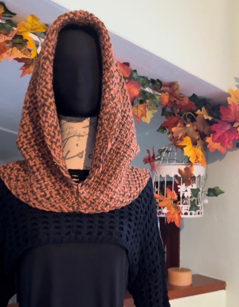 May include: A brown and orange knitted cowl with a hood. The cowl is displayed on a mannequin wearing a black top.