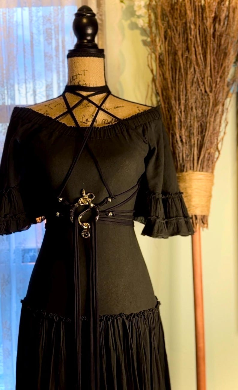 May include: A black off-the-shoulder dress with a gold snake-shaped belt and a black choker necklace. The dress has a ruffled hem and sleeves.