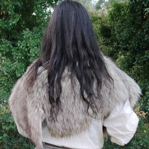 May include: A faux fur shawl with a light brown and white color pattern. The shawl is worn over a white shirt and a brown belt with a geometric pattern.