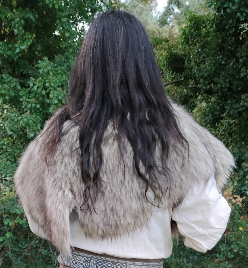 May include: A faux fur shawl with a light brown and white color pattern. The shawl is worn over a white shirt and a brown belt with a geometric pattern.
