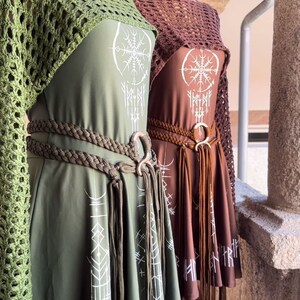 K&ouml;nnte beinhalten: Zwei gr&uuml;ne und braune Kleider mit wei&szlig;en geometrischen Mustern und Fransen. Die Kleider haben einen geflochtenen G&uuml;rtel mit einem gro&szlig;en Metallring.