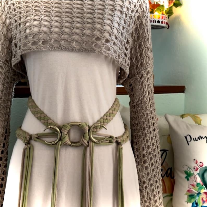 May include: A beige crochet top with long sleeves and a matching belt with a large, silver, crescent moon buckle. The belt has long, thin tassels.