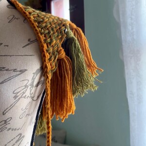 May include: A crocheted shoulder wrap with a woven pattern and tassels in shades of mustard yellow and olive green.
