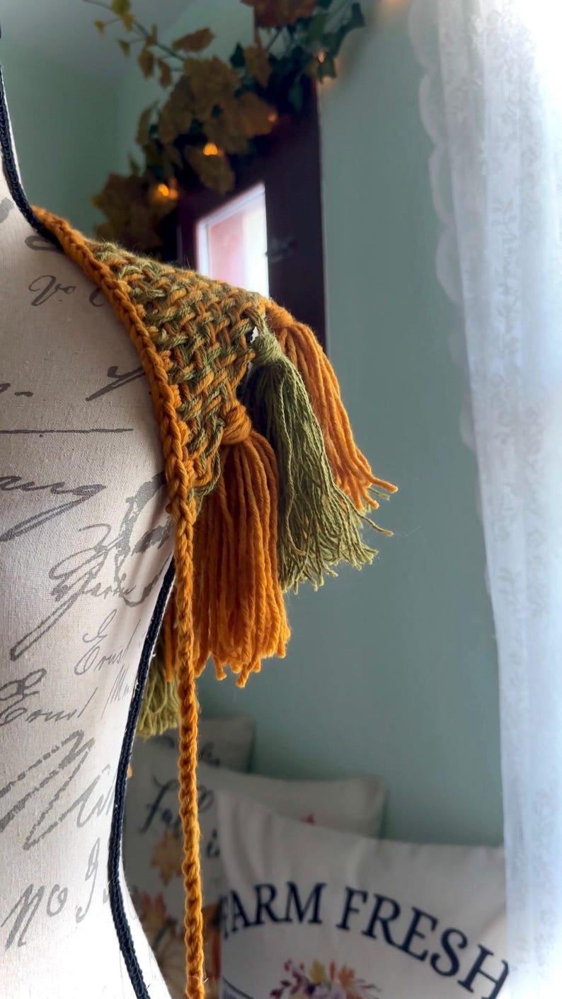 May include: A crocheted shoulder wrap with a woven pattern and tassels in shades of mustard yellow and olive green.