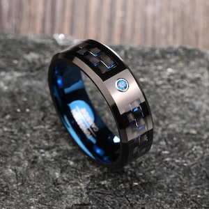 以下が含まれることがあります： 青いカーボンファイバーのインレイと小さな青い宝石が付いた、黒いタングステンカーバイドのリング。