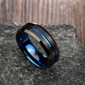 Puede incluir: Un anillo de carburo de tungsteno negro y azul con un borde biselado y una incrustación azul.