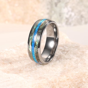 Op de afbeelding: Een zilverkleurige ring met een centrale band van blauwe opaal en iriserende abalone schelpinleg. De ring heeft een gepolijste afwerking en een afgerond profiel, met een mix van natuurlijke materialen en metallic glans.