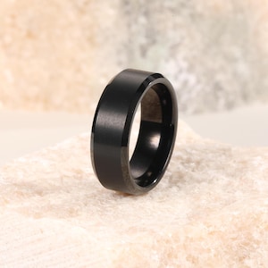 Könnte beinhalten: Ein schwarzer Ring mit gebürsteter Metalloberfläche. Der Ring hat eine flache Oberseite und abgeschrägte Kanten. Der Ring ist vor einem hellen, strukturierten Hintergrund platziert.