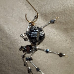 May include: A beaded spider ornament with a gold wire hanger. The spider is made of black and silver beads and has a round body and eight legs.