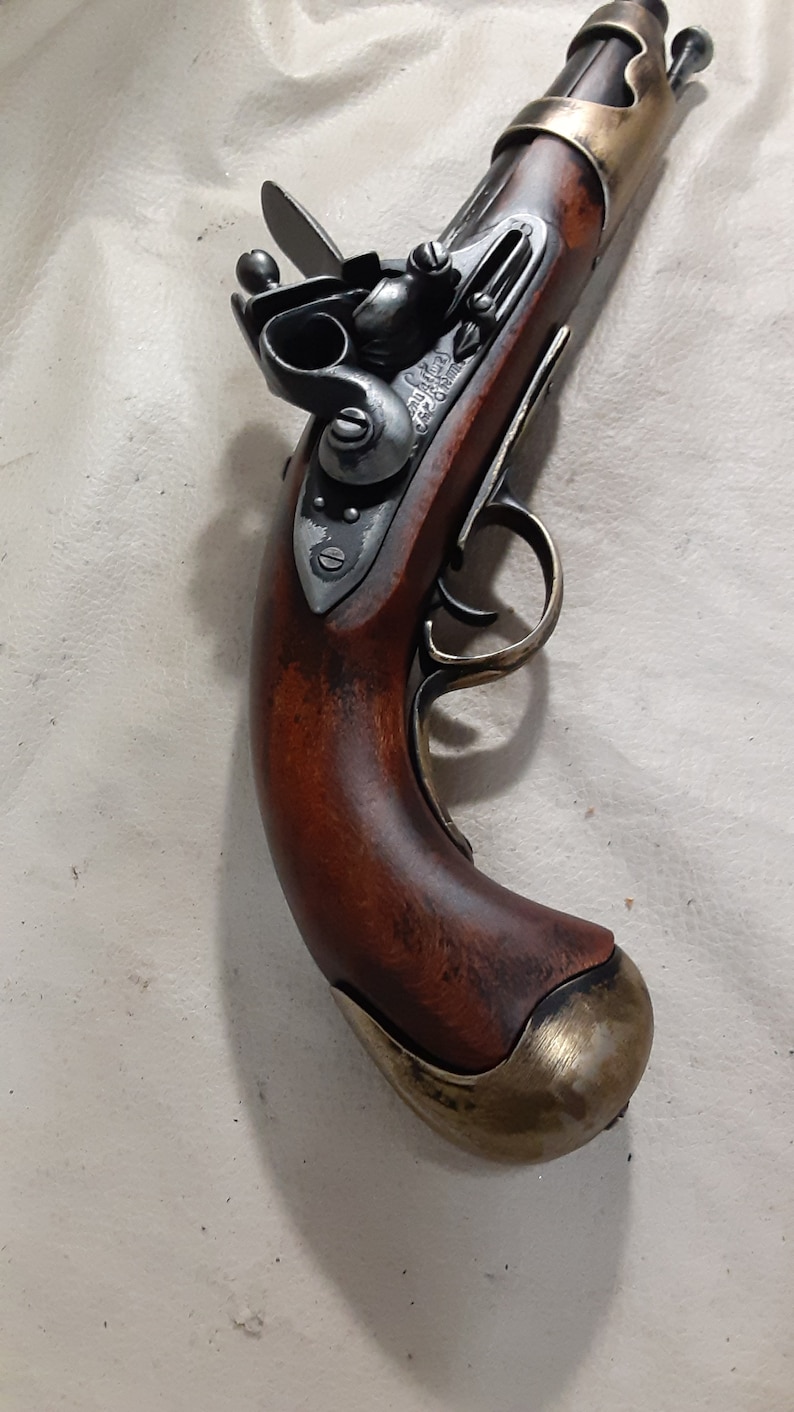 Op de afbeelding: Een antiek-stijl flintlock pistool met een houten handvat. Het pistool heeft een messing loopband en een messing trekkerbeugel. Het pistool is in goede staat en heeft een donkerbruine afwerking.