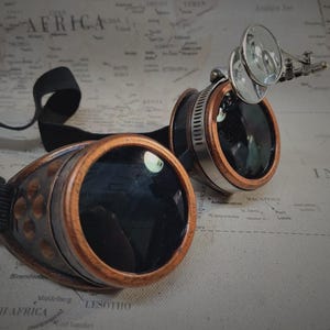 May include: A pair of steampunk goggles with brown wooden frames and black lenses. The goggles have a silver metal band and a magnifying glass attached to the side.