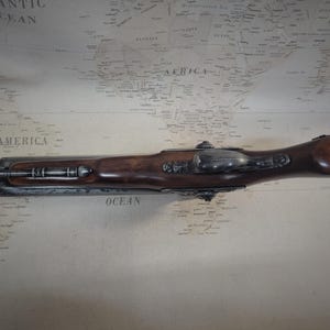 May include: An antique flintlock pistol with a black and silver finish and a wooden handle. The pistol is lying on a map with the words "South America" and "Atlantic Ocean" visible.