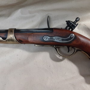 Op de afbeelding: Een zwart en messing vuursteenpistool met een houten handvat. Het pistool heeft een messing trekkerbeschermer en een messing loopband. Het pistool is gegraveerd met de tekst "J.M.  LONDON  260".