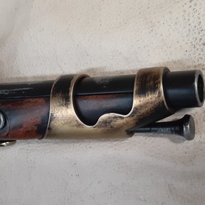 Op de afbeelding: Een zwart en bruin flintlock-pistool met een gouden metalen loopband. Het pistool heeft een houten kolf en een metalen hamer.