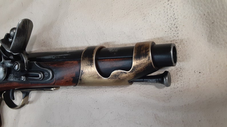 Op de afbeelding: Een zwart en bruin flintlock-pistool met een gouden metalen loopband. Het pistool heeft een houten kolf en een metalen hamer.