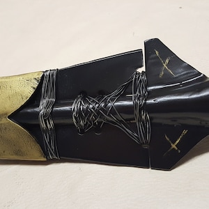 May include: A black and gold spearhead with a braided cord wrapped around the shaft. The spearhead has a pointed tip and a flat, triangular blade. The cord is tied in a knot at the base of the blade.
