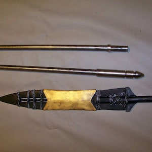 May include: A gold-colored spearhead with a black metal shaft and a black and silver braided design. The spearhead is attached to a long, silver metal shaft.