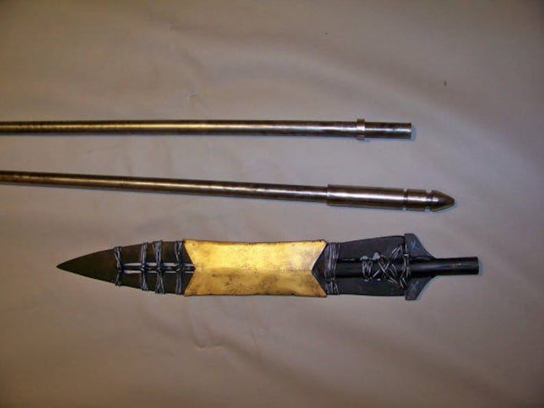 May include: A gold-colored spearhead with a black metal shaft and a black and silver braided design. The spearhead is attached to a long, silver metal shaft.