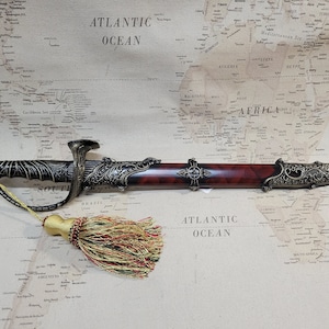 May include: A decorative dagger with a brown wooden handle and ornate gold metal accents. The dagger has a blue gemstone at the tip and a tassel hanging from the handle.
