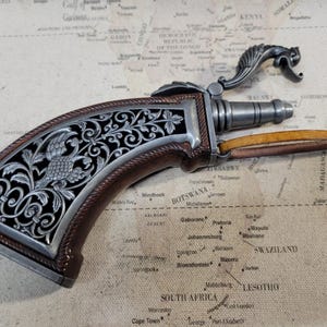 May include: A decorative, ornate, silver-coloured pistol with a brown wooden handle. The pistol has intricate floral designs and a small bird on the handle. The pistol is lying on a map.