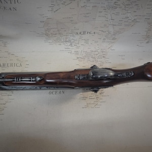 May include: A black and silver antique flintlock pistol with a wooden handle. The pistol is lying on a map with the words "South America" and "Atlantic Ocean" visible.