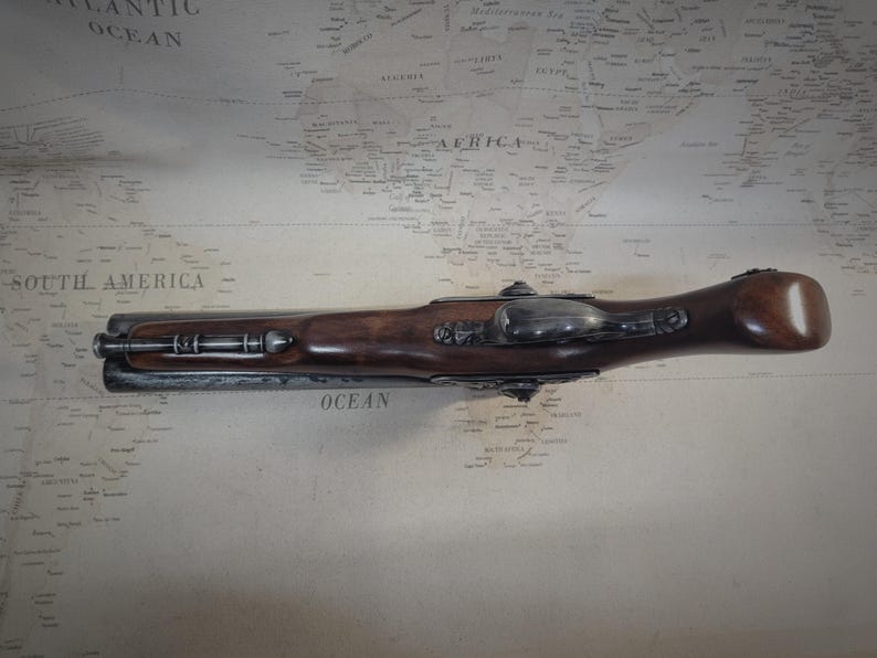 May include: A black and silver antique flintlock pistol with a wooden handle. The pistol is lying on a map with the words "South America" and "Atlantic Ocean" visible.