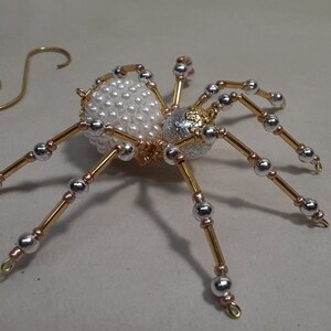 May include: A decorative spider ornament with a body covered in white pearls and a silver and gold head. The spider has gold and silver legs with silver beads. A gold wire spiral is next to the spider.