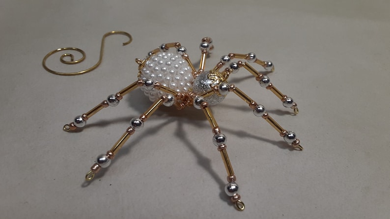 May include: A decorative spider ornament with a body covered in white pearls and a silver and gold head. The spider has gold and silver legs with silver beads. A gold wire spiral is next to the spider.
